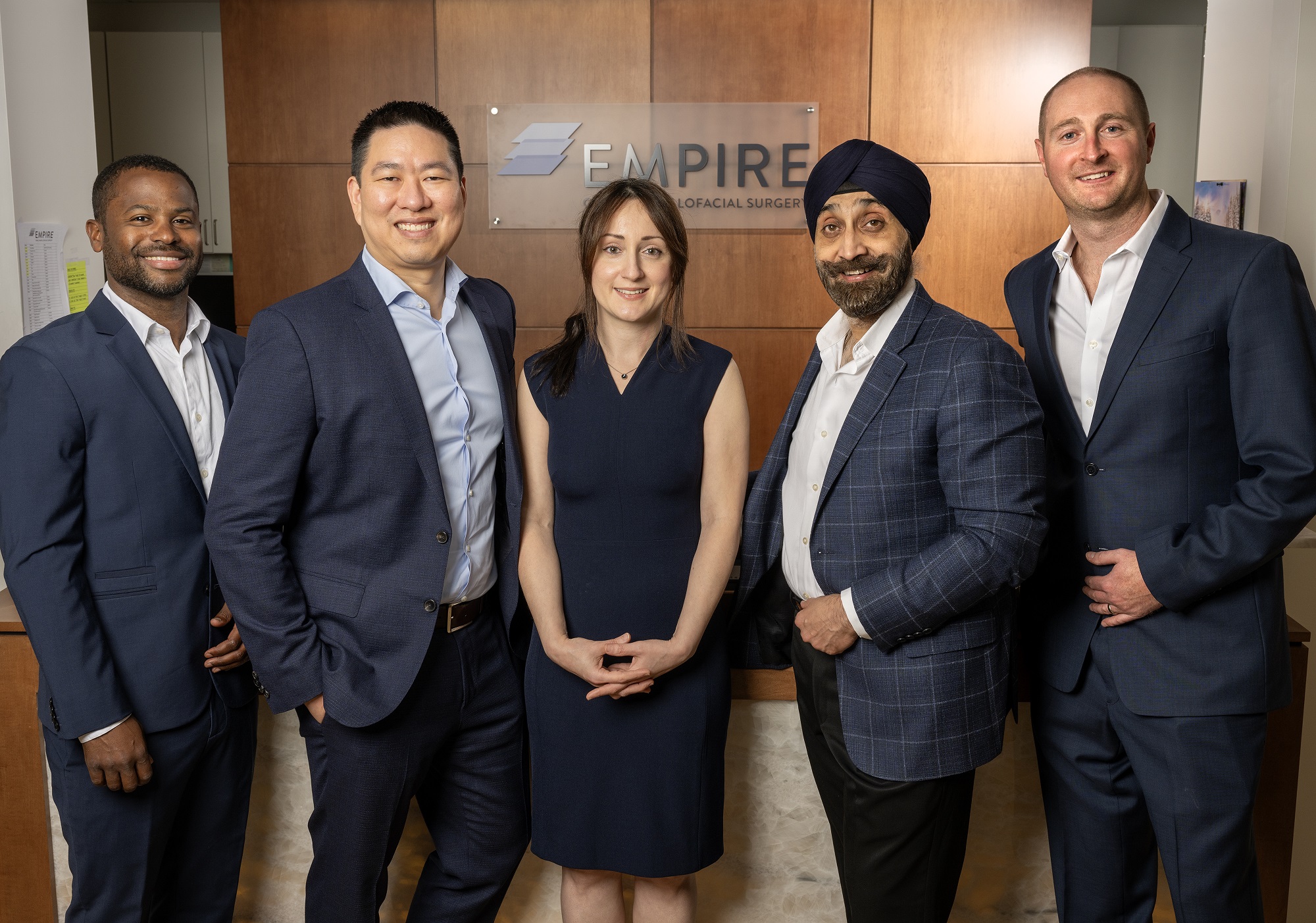 Dr. Johar, Dr. Adams, Dr. Boliere, Dr. Barzani and Dr. Wun smiling standing together in front of the Empire Oral and Maxillofacial sign in the office