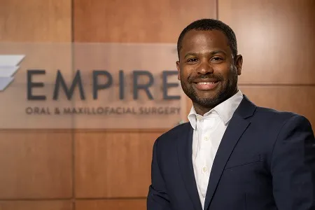 Dr. Boliere smiling in front of the Empire Oral and Maxillofacial Surgery sign in the office